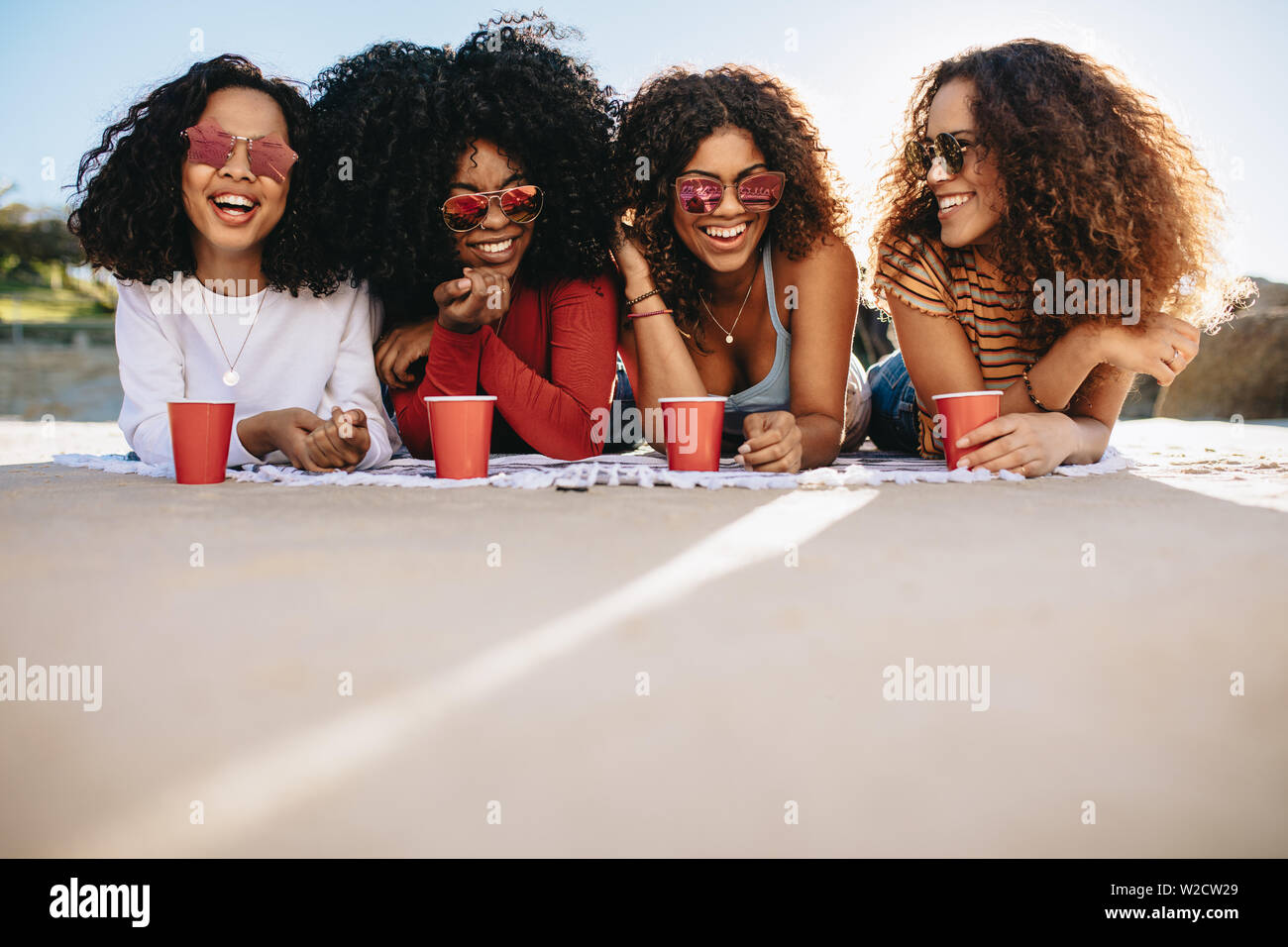 Gruppe von Mädchen mit lockigem Haar liegen am Strand mit kalten Getränken. Schöne Frauen entspannen am Strand am Wochenende. Stockfoto