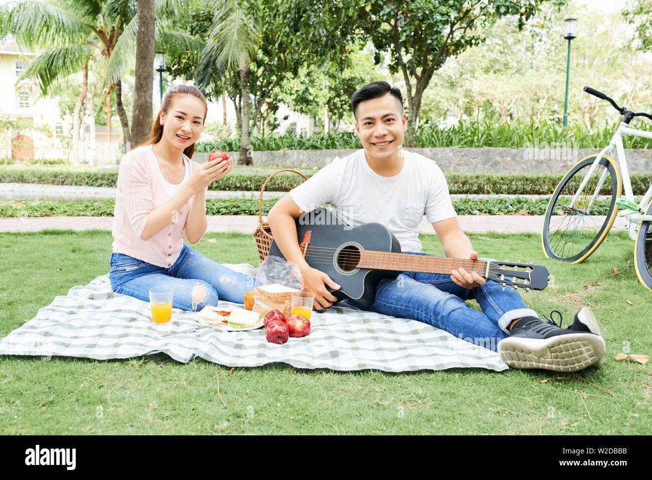 Portrait von Asiatischen junges paar Lächeln auf die Kamera beim Sitzen auf dem Rasen Obst essen und spielen auf der Gitarre im Sommer Picknick im Park Stockfoto