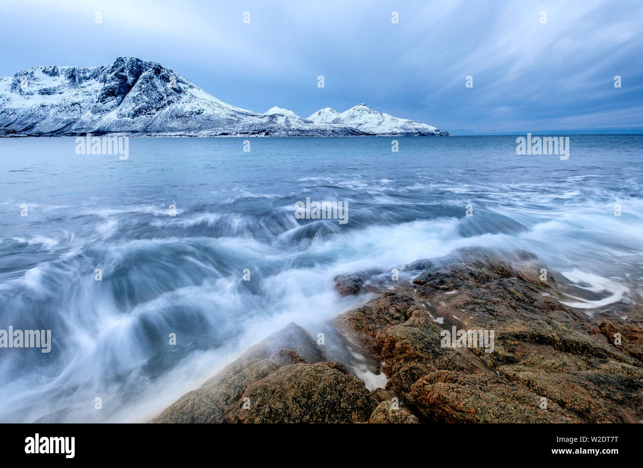 Schwere Wellen brechen auf braunen Felsen mit schneebedeckte Berg im Hintergrund, Norwegen Stockfoto