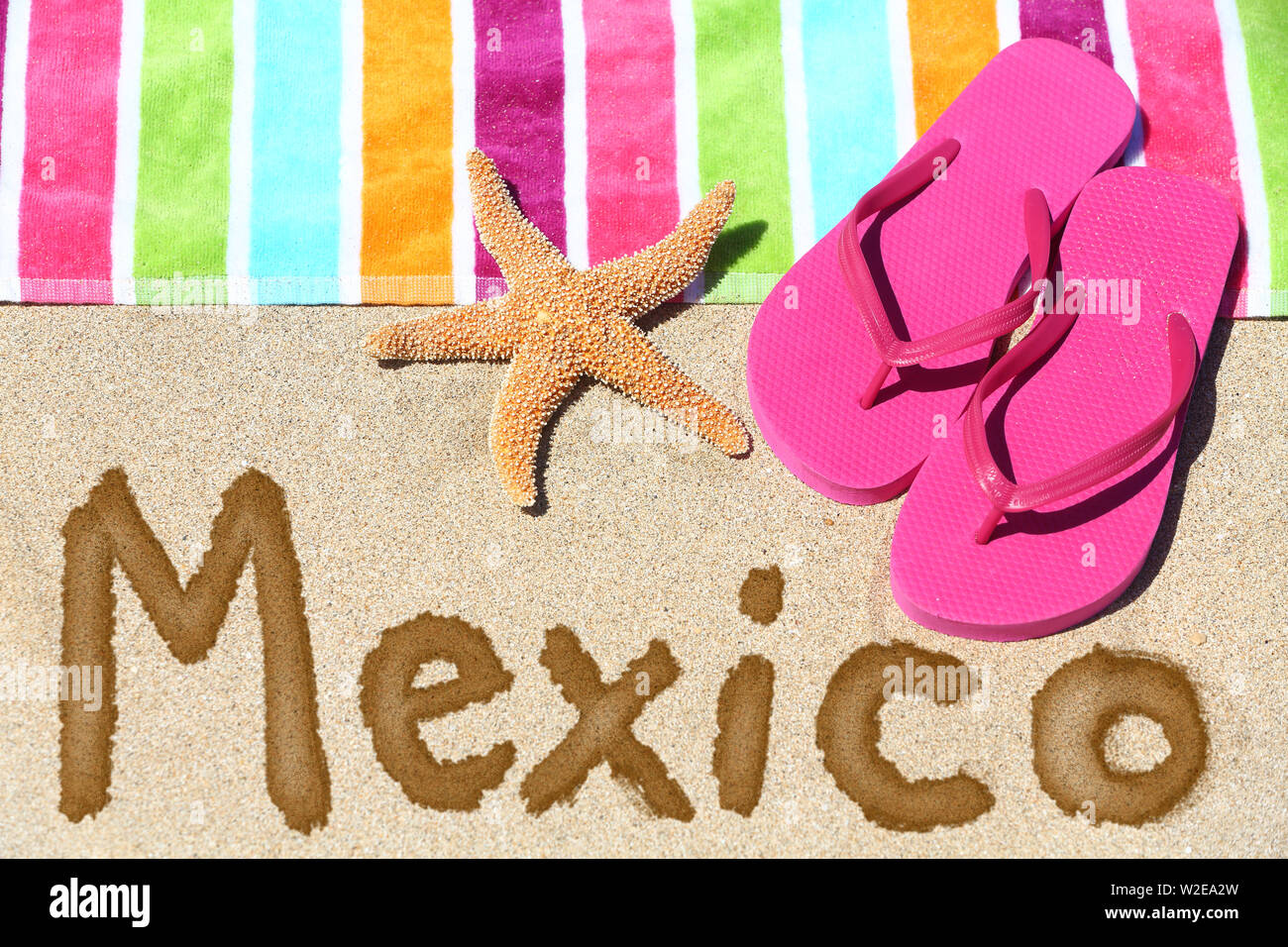 Mexiko Strand Travel Concept. Mexiko im Sand mit Wasser neben Handtuch für den Strand und Sommer Sandalen und Seesterne geschrieben. Mexikanische Ferien Urlaub Foto. Stockfoto