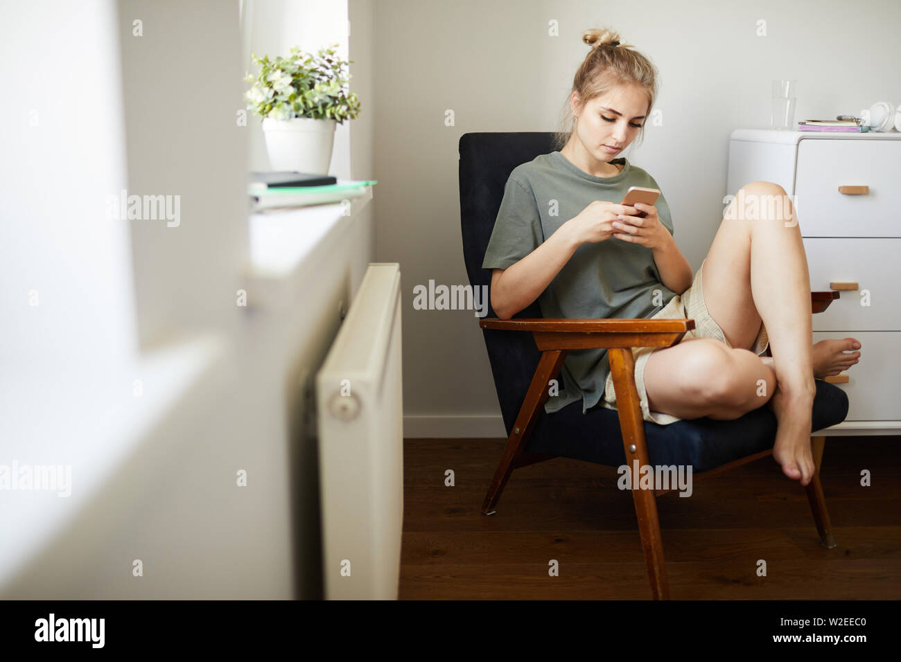 Süße Mädchen in casualwear Video im Smartphone beim Entspannen im Sessel im Schlafzimmer Stockfoto