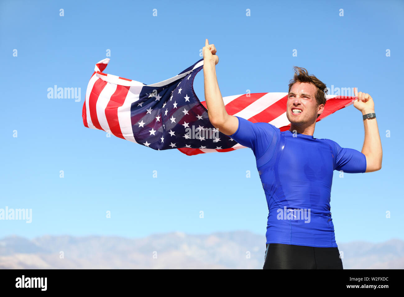 Athleten zujubeln Holding US Flag kompromisslos sportlichen jungen Mann, der mit der amerikanischen Flagge in die Luft hob in seinen Händen weht im Wind gegen einen klaren sonnigen blauen Himmel Stockfoto