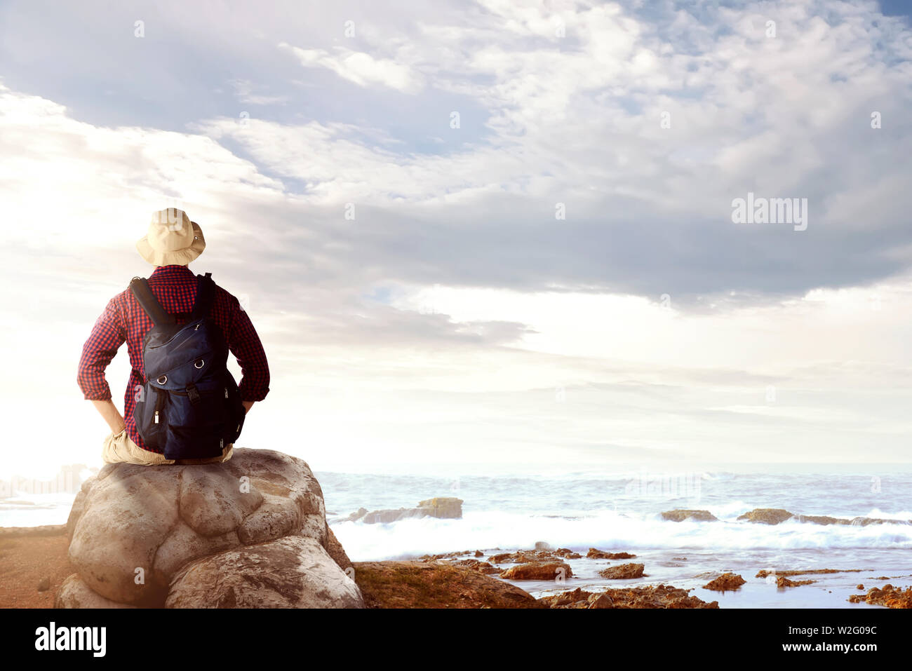 Ansicht der Rückseite des asiatischen Mann in Hut mit Rucksack sitzen auf den Felsen und mit Blick auf das Meer Blick auf den Strand Stockfoto