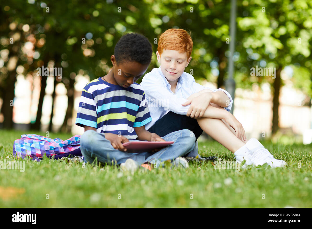 Afro-amerikanische Teenager Schüler sitzen auf Gras spielen mit Tablette während sein Freund beobachten Stockfoto