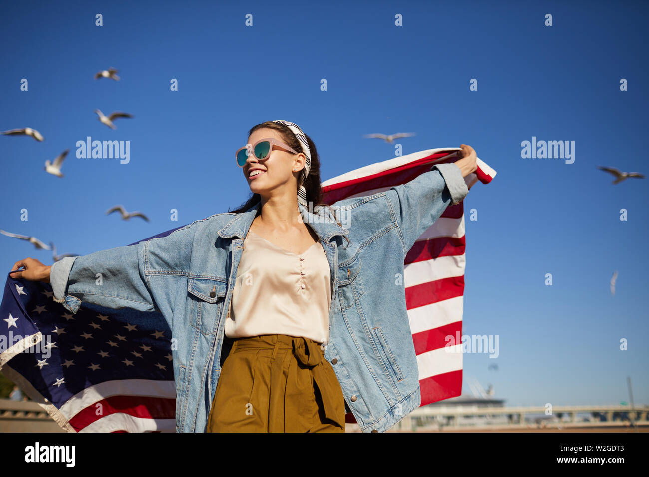 Happy Independent girl in modischen Sonnenbrillen und Jeansjacke outstretching Arme und die amerikanische Flagge, Möwen im Hintergrund fliegen Stockfoto