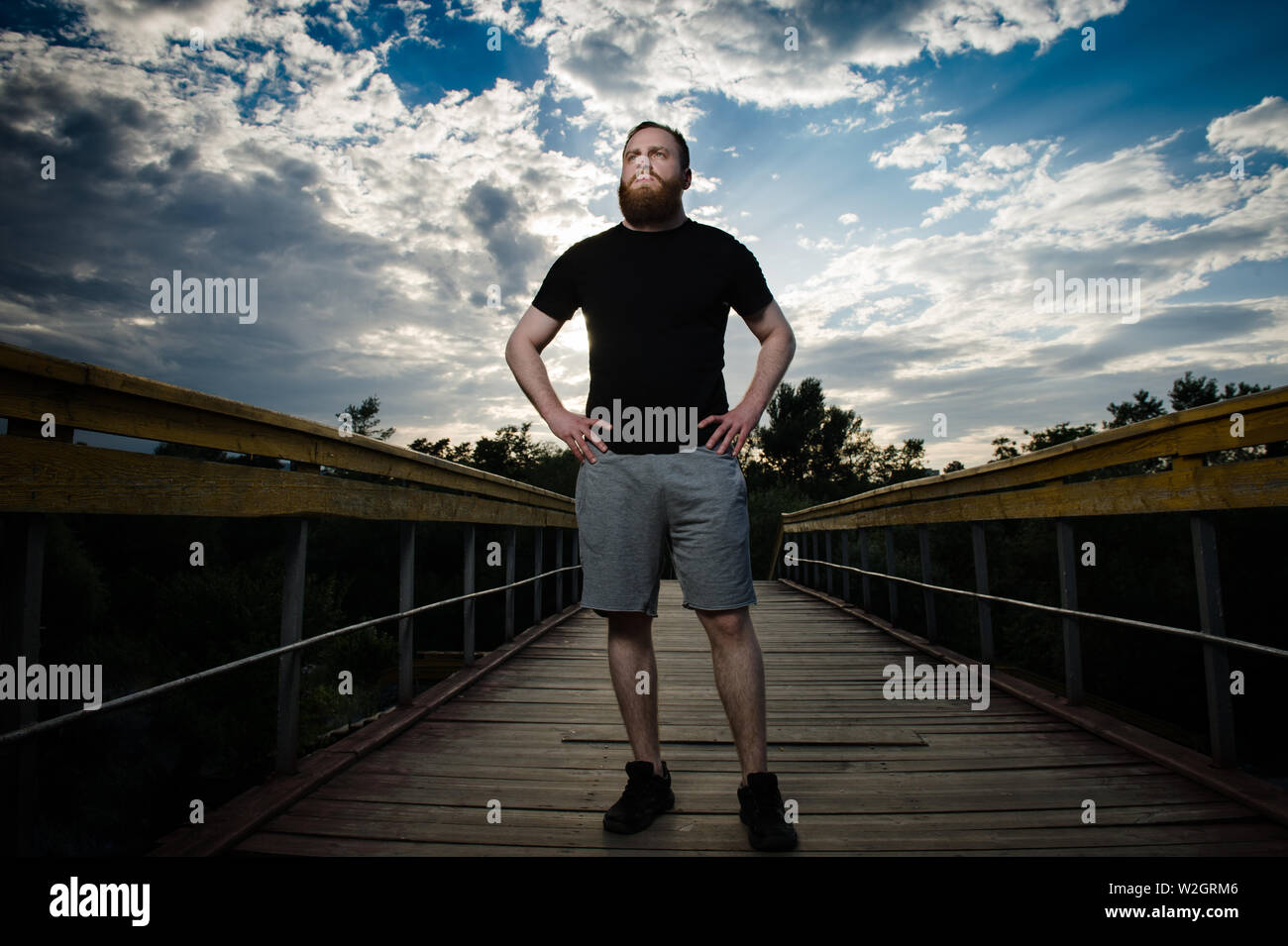 Dramatisches Bild der starken jungen Mann mit Bart in schwarzen T-Shirt und graue Shorts stehen auf Holz Brücke im Park mit Sonnenuntergang Himmel mit Wolken Stockfoto