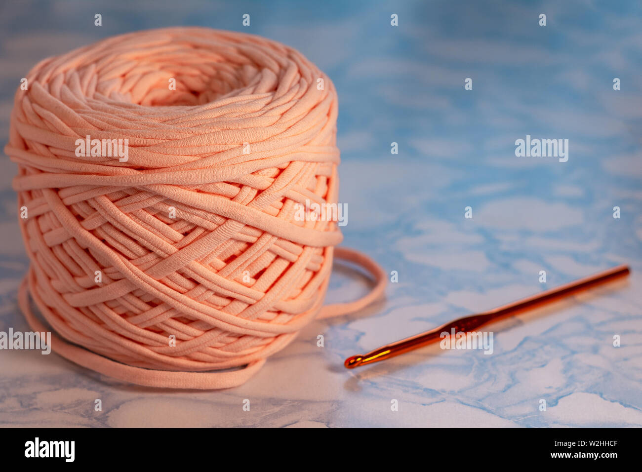 Hobbys und kreative Aktivitäten Konzept - eine Spule von großen Gestrickte Garne aus Pfirsich Farbe mit orangefarbenen glänzenden Haken auf der Oberfläche der blauen Marmor. Stockfoto