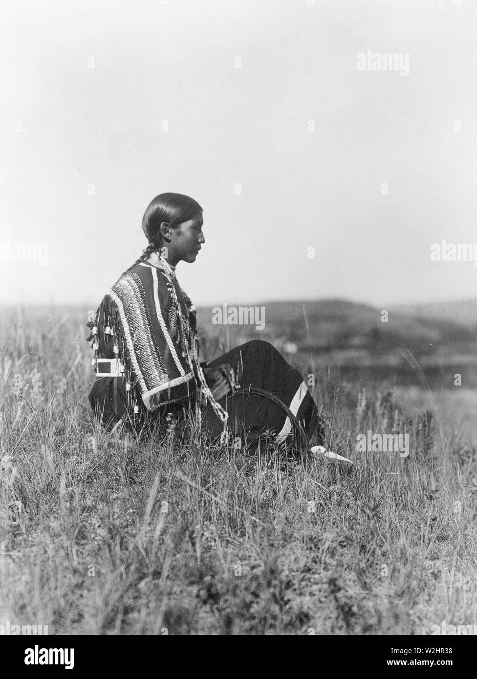 Edward S. Curtis Native American Indians - Junge Piegan indische Frau, tragen Perlen Kleid, auf einem Hügel sitzen kann. 1910 Stockfoto