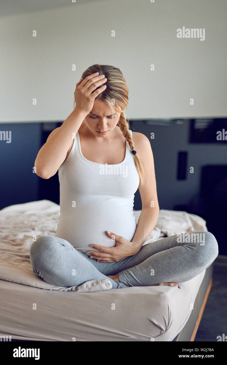 Stark gestörten schwangere junge Frau sitzt im Schneidersitz auf einem Bett mit ihrem aufgeblähten Bauch und hielt den Kopf gesenkt, mit einem ernsten Ex Stockfoto