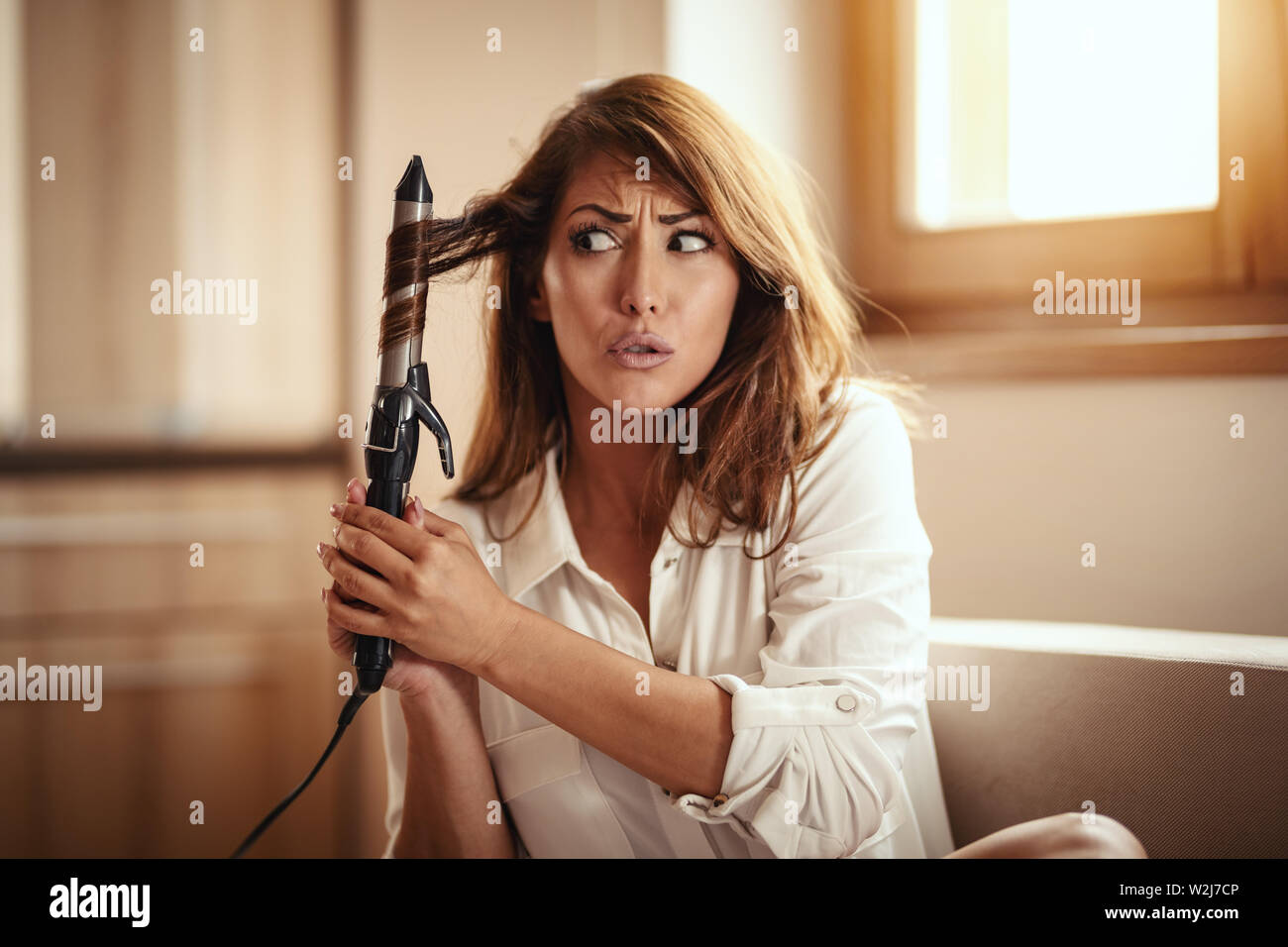 Eine junge Frau besorgt locken ihr Haar mit einem elektrischen Lockenwickler und weiß nicht, ob die Frisur sehr gut. Stockfoto