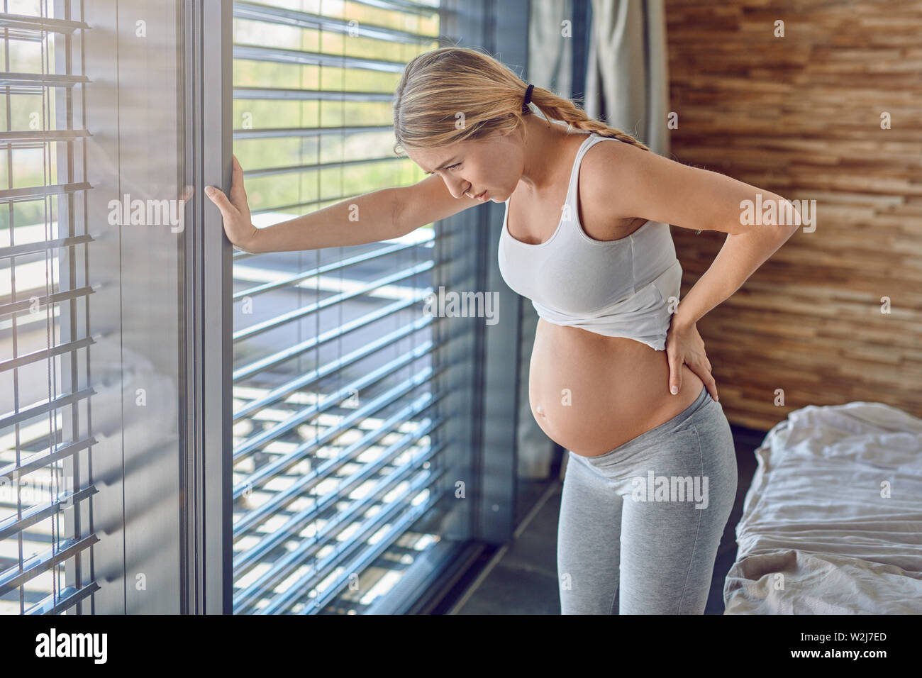 Schwangere junge Frau leiden mit Rückenschmerzen lehnte sich auf ihr Bett, Ihr unterer Rücken umklammert Mit einem gequälten Ausdruck Stockfoto