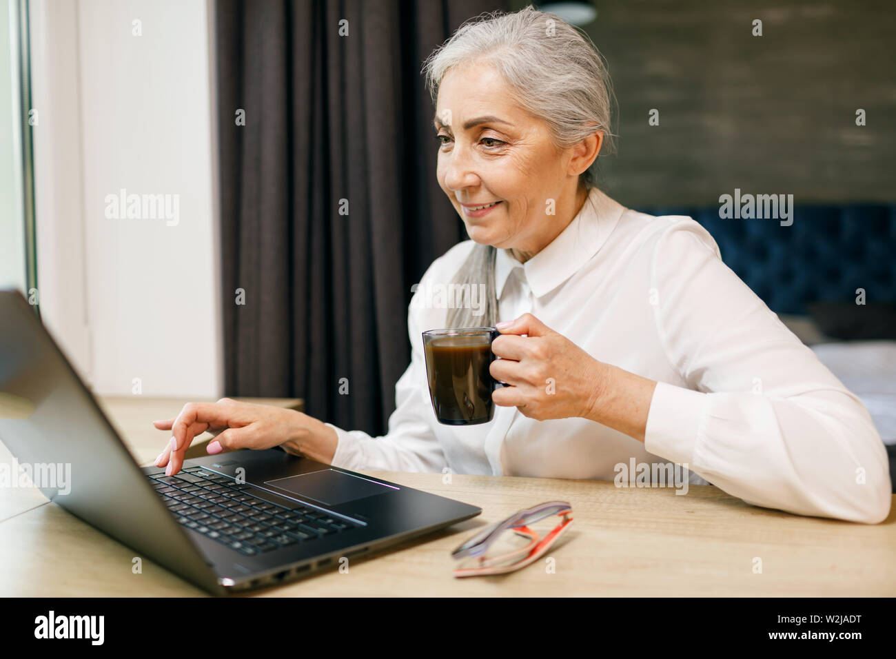 Ältere Frau mit weißen Haaren trinken Kaffee während der Arbeit mit dem Laptop Stockfoto