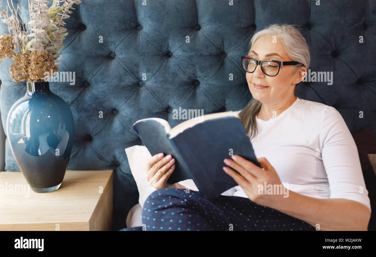 Ziemlich alte Frau mit dem grauen Haar in Brillen Buch in Schlafzimmer Stockfoto