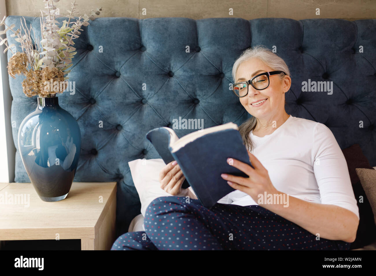 Lächelnde ältere Frau mit grauen Haaren in Brillen Festlegung auf dem Bett und lesen Buch im Schlafzimmer Stockfoto