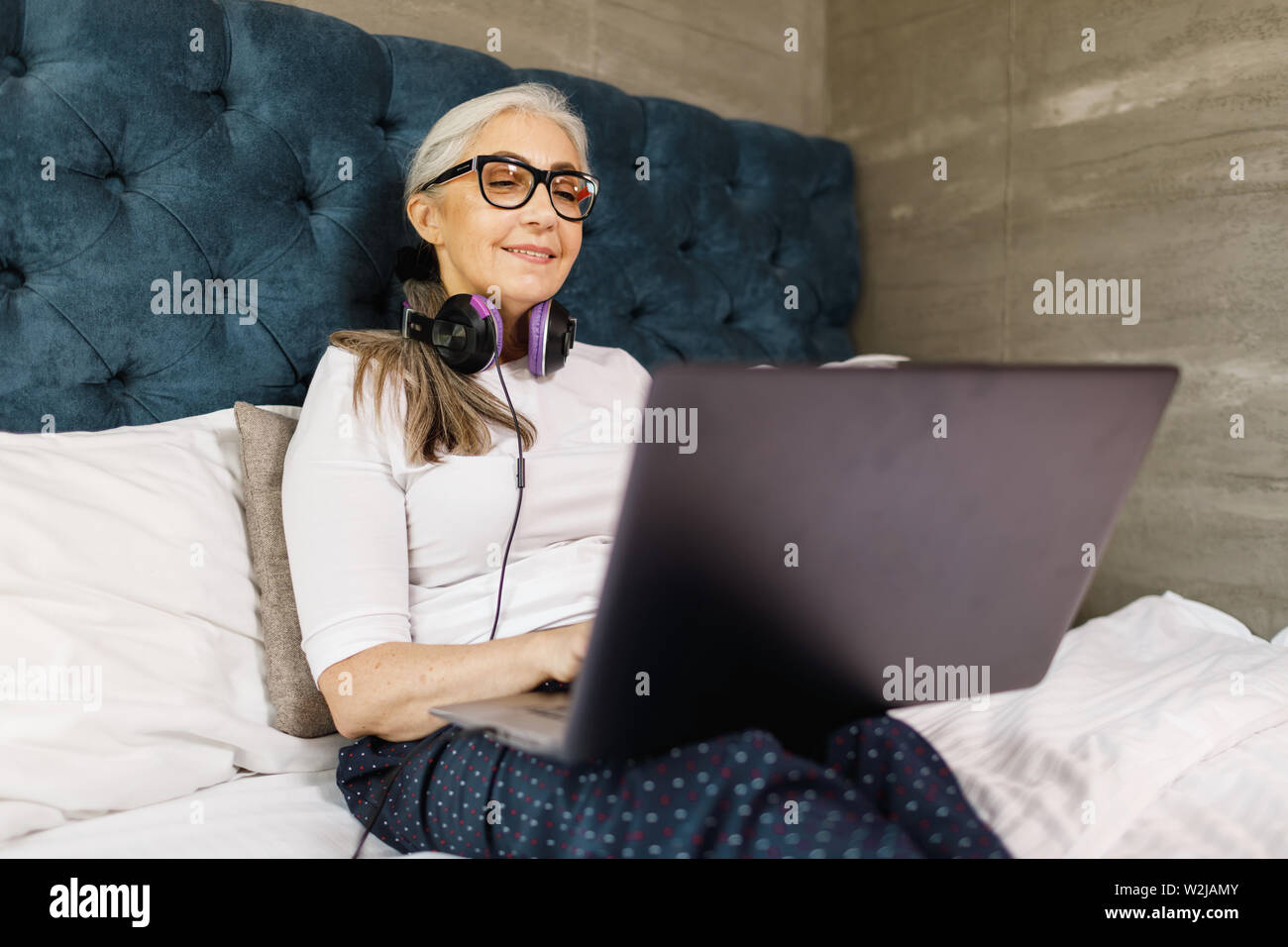 Ältere Frau mit weißen Haaren in Brillen Tragen von Kopfhörern watching Film auf dem Laptop bei der Verlegung auf dem Bett Stockfoto