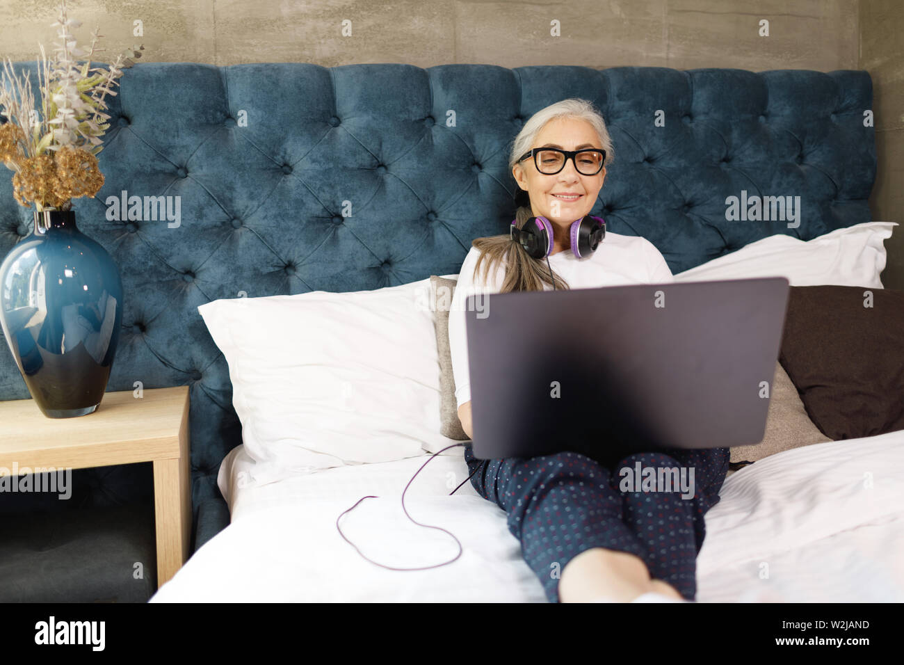 Alte Frau mit dem grauen Haar in Brillen Tragen von Kopfhörern Festlegung auf dem Bett mit Laptop Stockfoto