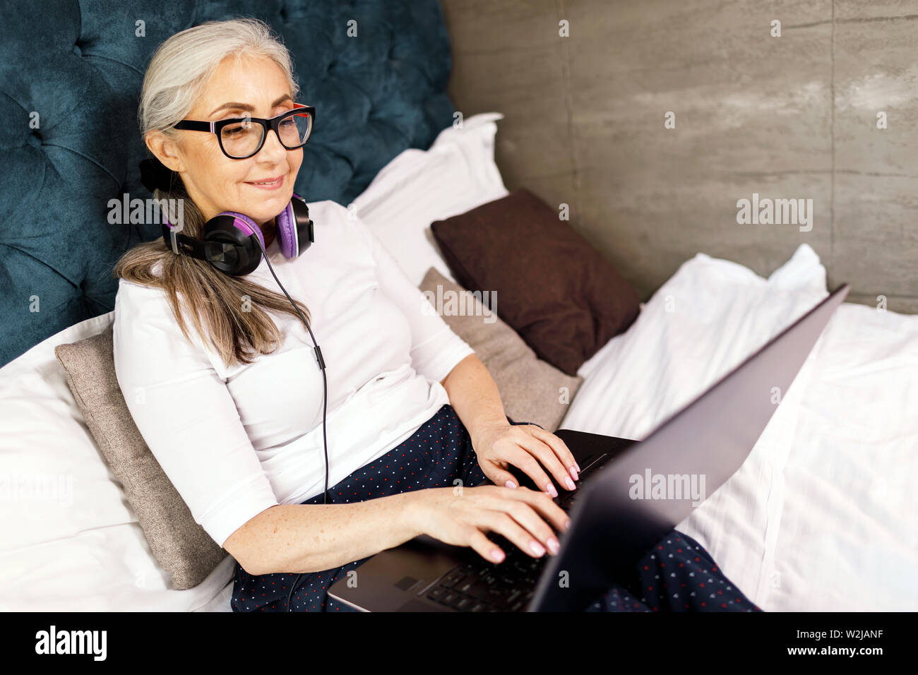 Ältere Frau mit weißen Haaren in Brillen Tragen von Kopfhörern plaudern auf dem Laptop bei der Verlegung auf dem Bett Stockfoto