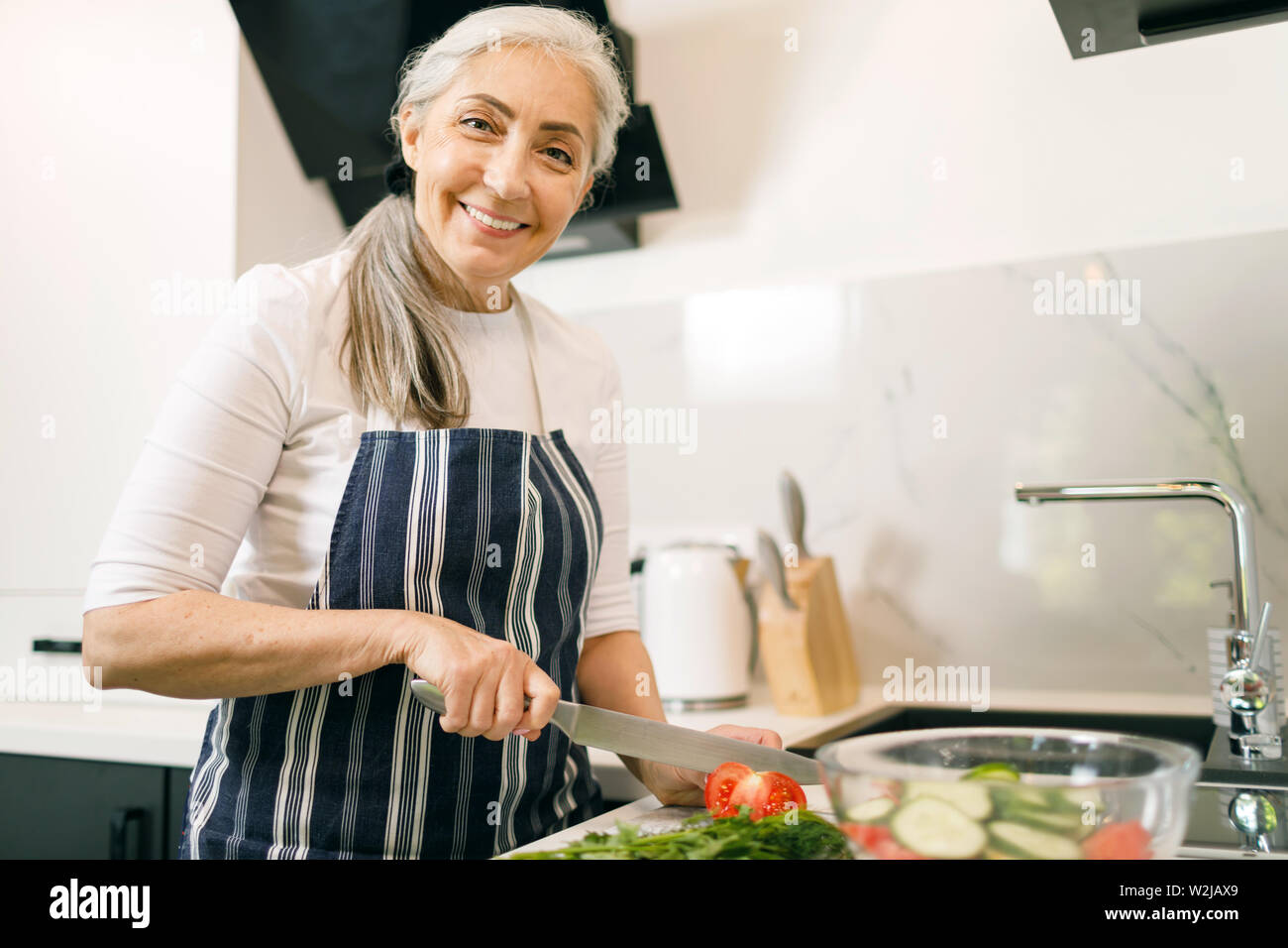 Lächelnde ältere Frau mit weißen Haaren in eine Schürze Schneiden von Gemüse auf einer Holzplatte in Ihrer Küche Stockfoto