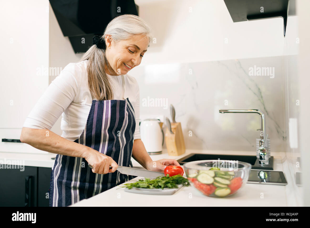 Lächelnde ältere Frau mit grauen Haaren im Vorfeld vorbereiten Salat in der Küche Stockfoto