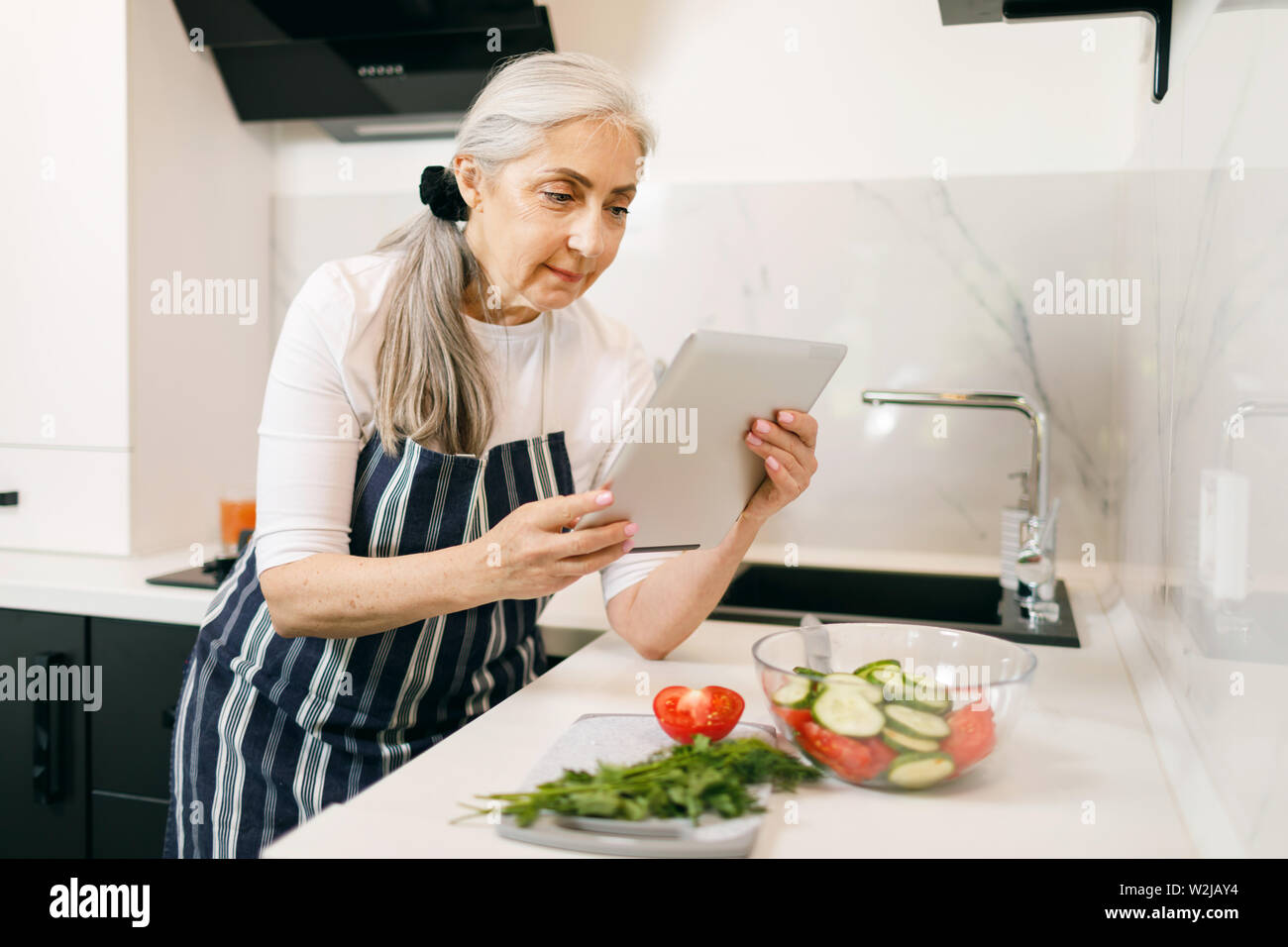 Lächelnde ältere Frau mit weißen Haaren in eine Schürze beobachten Rezepte an die Tablette beim Kochen in der Küche Stockfoto
