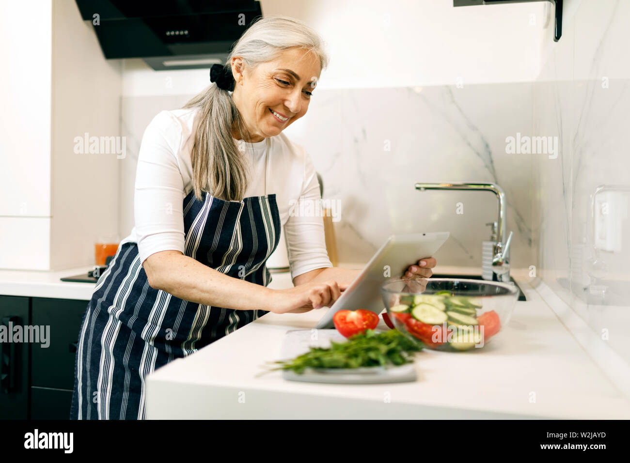 Lächelnde ältere Frau mit weißen Haaren in eine Schürze mit Tablette beim Kochen in der Küche Stockfoto