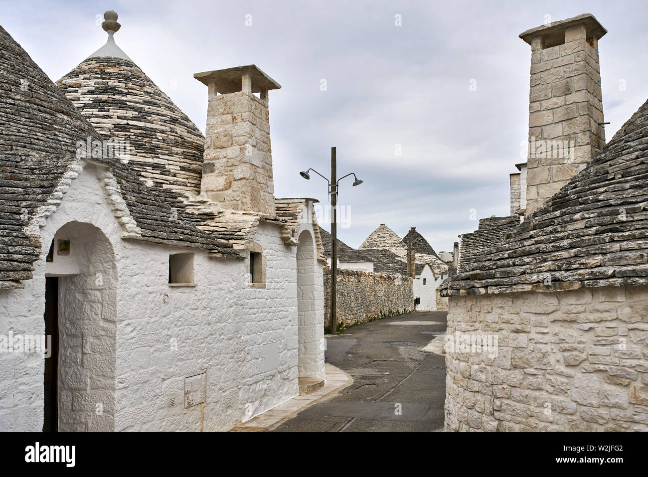 Mittelalterliche Straße mit Trulli auf dem bewölkten Himmel Hintergrund in Alberobello Stadt in Italien. Es gibt große Kamine auf den Dächern. Horizontale. Stockfoto