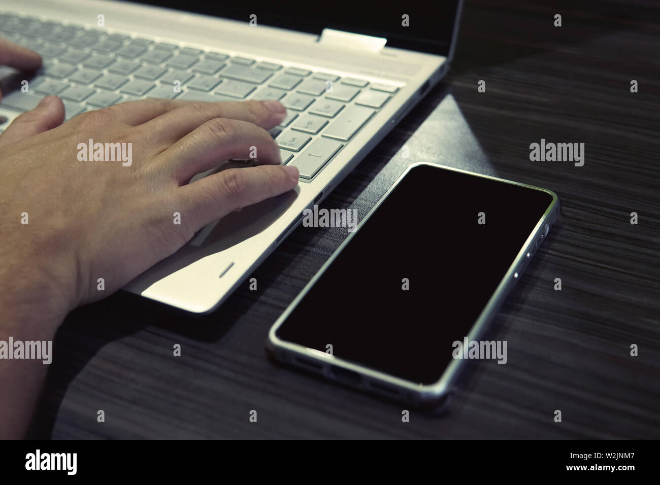 Mann hinter einem Laptop und Smartphone, Hände auf der Tastatur sitzen. Schwarz - aussortiert Telefon ist am Tisch in der Nähe des Notebooks mit männlichen Händen auf der Tastatur Stockfoto