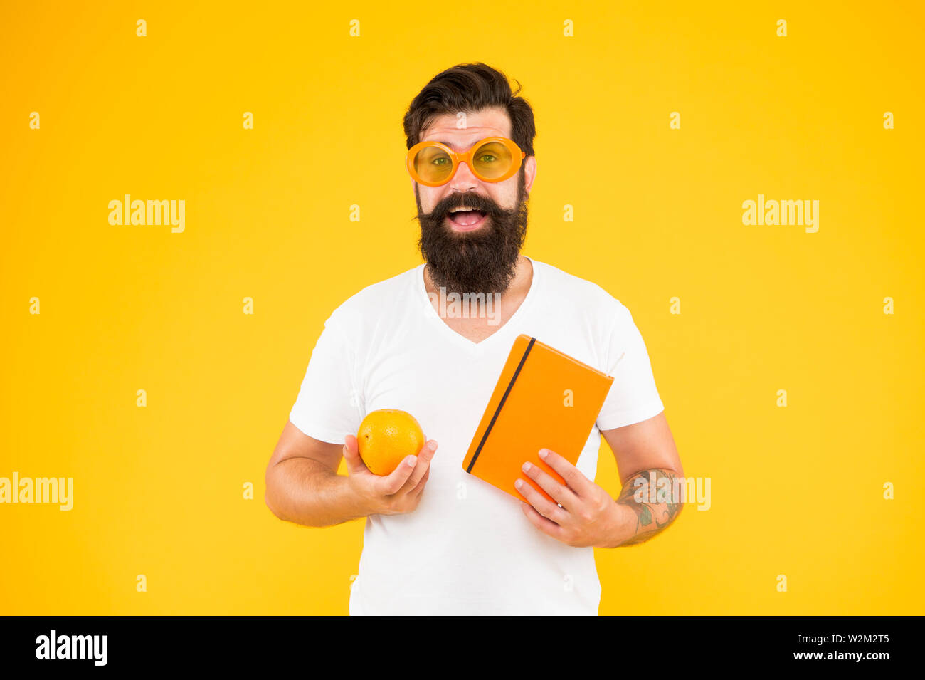 Stolz der geek in Sie. Geek Mann. Bärtiger Mann in geek Brille Holding orange und Buch auf gelben Hintergrund. Hipster in Geek Chic style Auswahl gesunder Nahrung für das Gehirn. Stockfoto