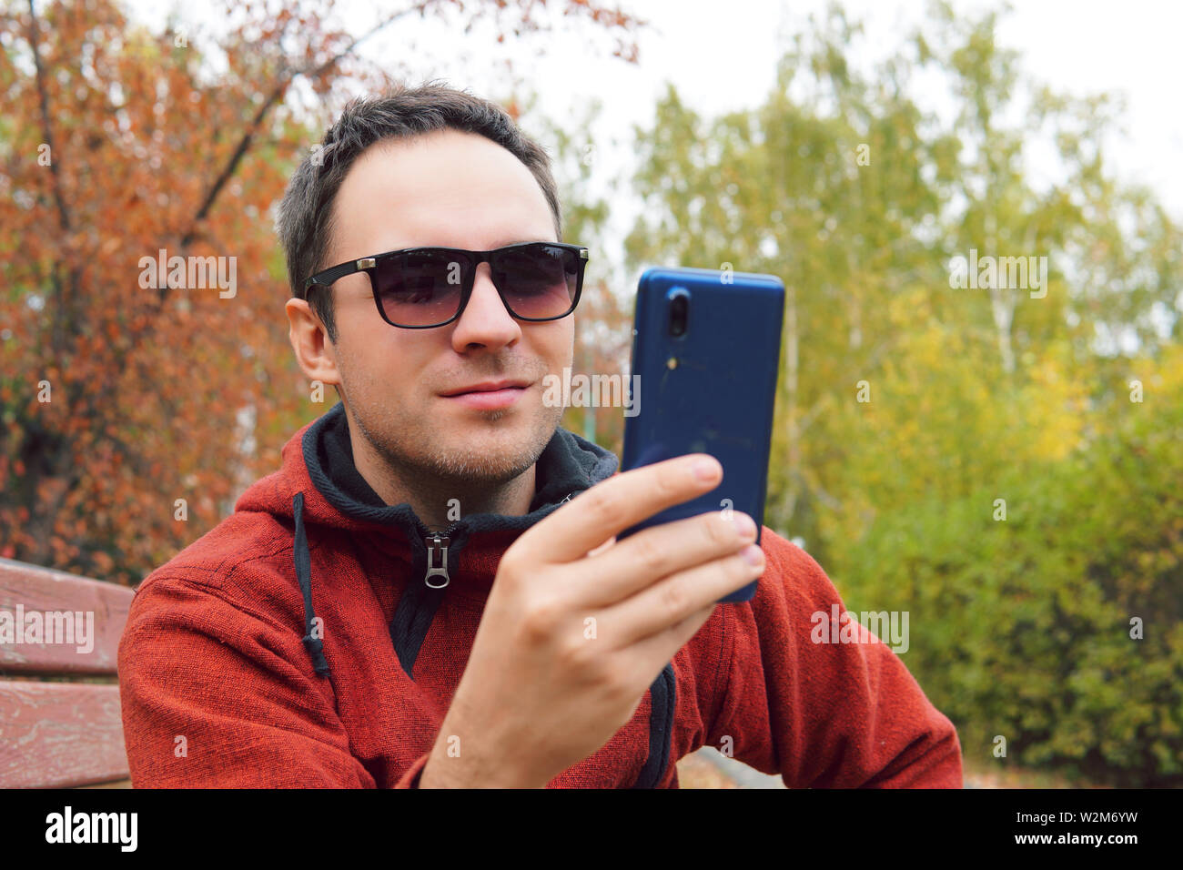 Europäische Kerl sitzt auf einer Bank in einer Stadt Herbst Park. Ein Mann ist ein Lächeln am Telefon. Konzept der Freude und Vergnügen, Freude und Lachen. Humor Stockfoto