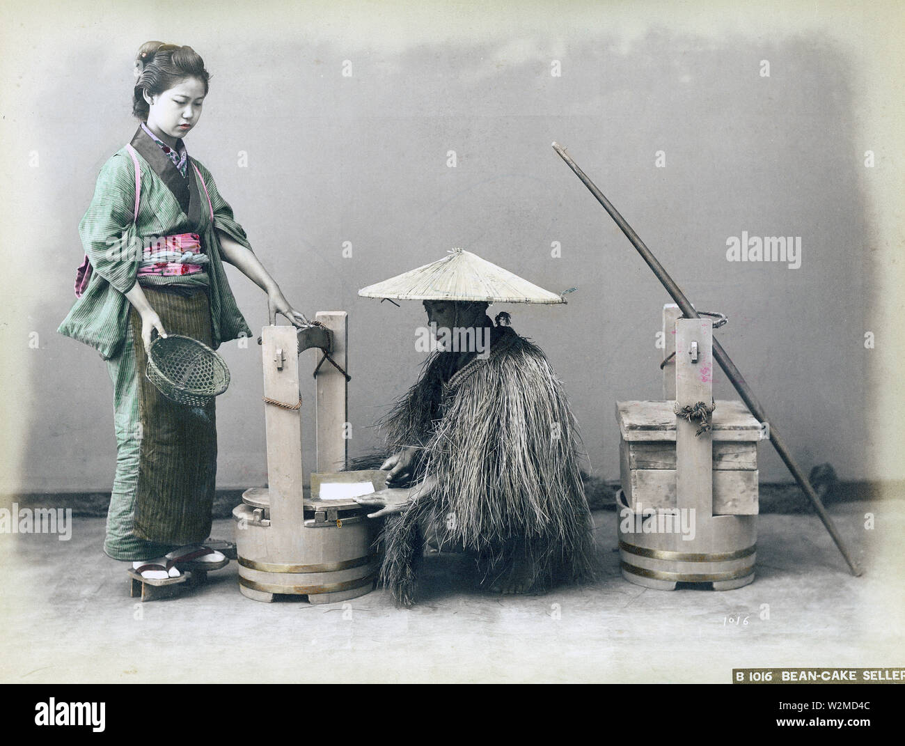 [1880s Japan - Japanische Tofu Anbieter] - ein Tofu (Tofu) Hersteller tragen eines Mino (Stroh regenmantel) und einem sugegasa Konische hat Schichten einen Block von Tofu mit einem Messer. Eine Frau im Kimono und traditionelle Frisur hält einen Korb. Eine sehr ökologische Weise Tofu zu verkaufen! 19 Vintage albumen Foto. Stockfoto