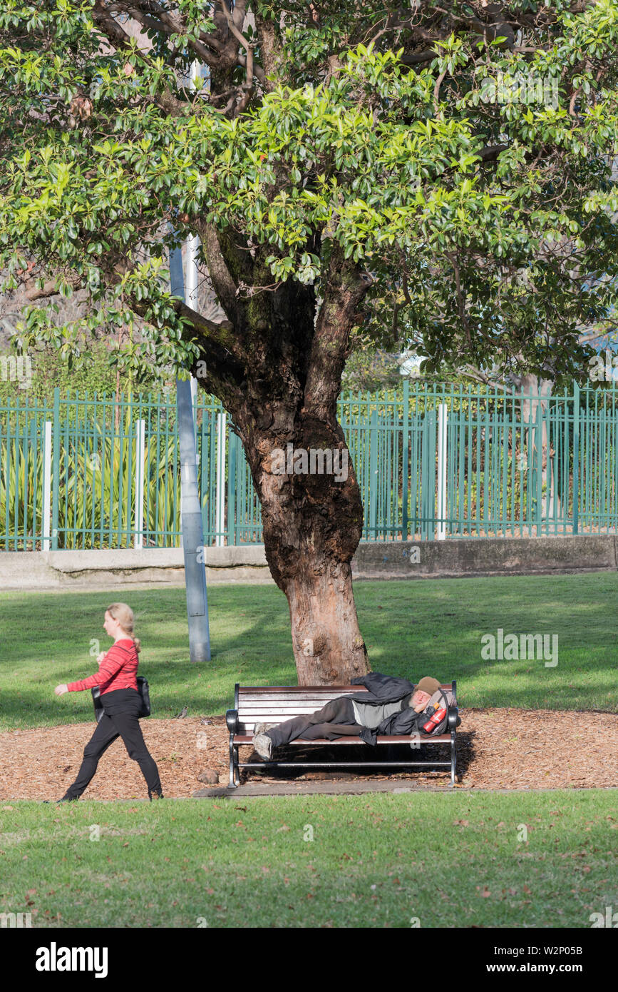 Eine Frau, die an einem obdachlosen älteren Mann vorbeigeht, der auf einer Parkbank in einem Stadtpark in Sydney in Australien schläft Stockfoto