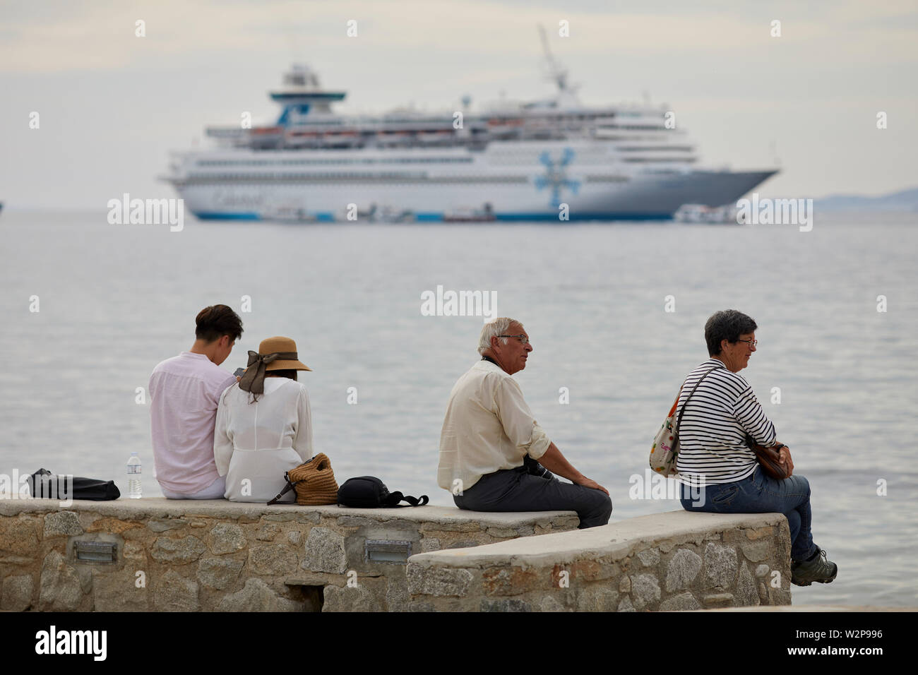 Mykonos, griechische Insel Mikonos, Teil der Kykladen, Griechenland. Touristen auf a waterfront Beach Bank auf der Suche nach einem brüllte Kreuzfahrtschiff im Hafen Stockfoto