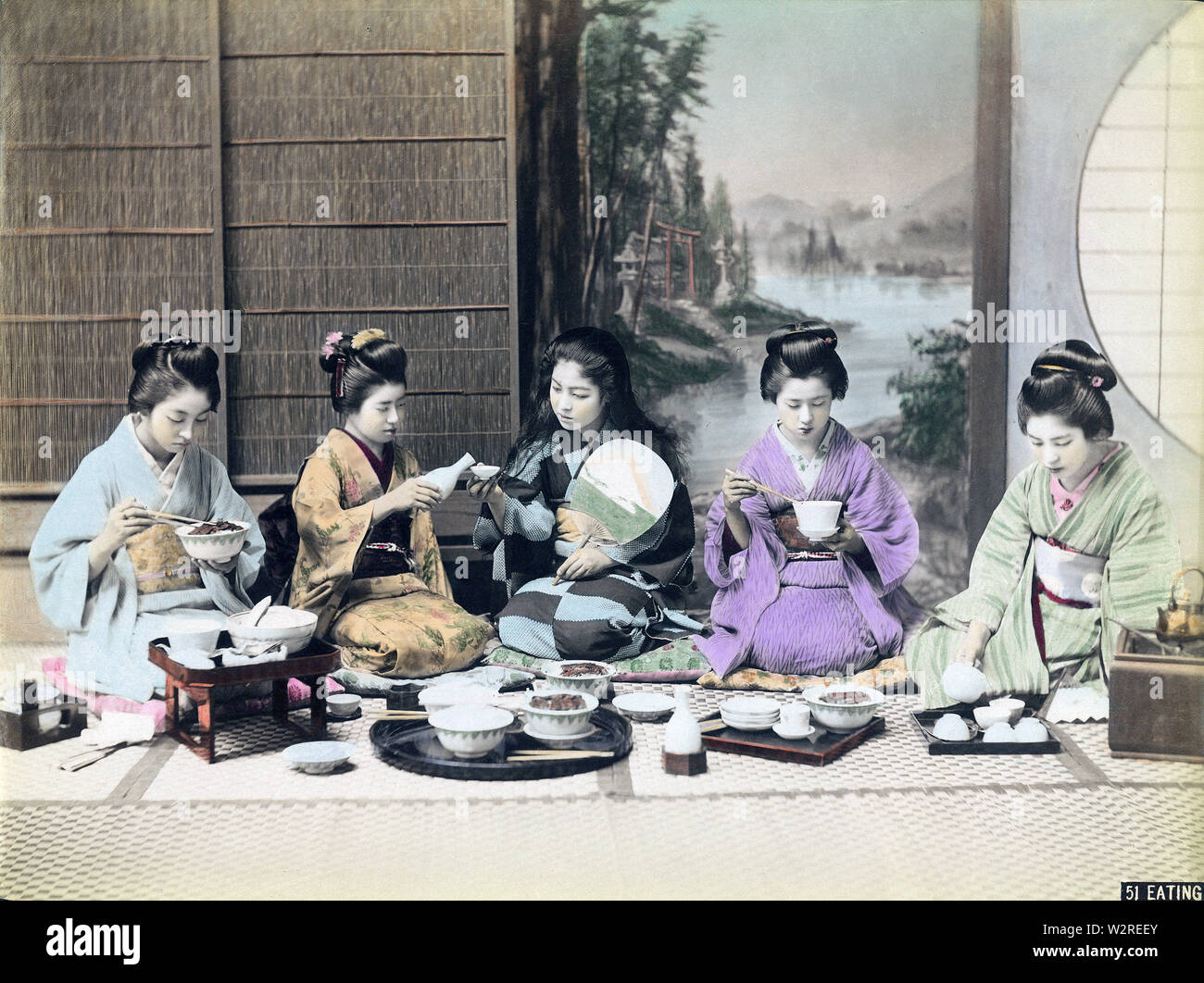 [1890s Japan - Frauen in einer Mahlzeit] - in einer Mahlzeit. Fünf junge Frauen im Kimono auf dem Boden sitzt. Zwei Frauen sind mit Stäbchen, Man gießt Sake (Reiswein), in die Schale der Frau neben ihr, während die fünfte Gießen ist Tee. 19 Vintage albumen Foto. Stockfoto