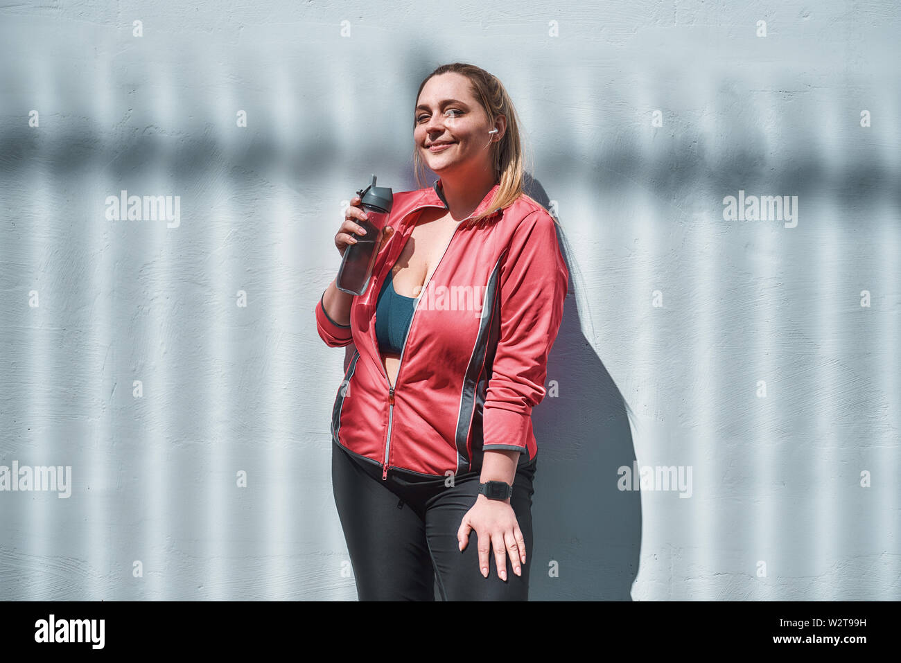 Ich liebe meinen Körper Junge und positive plus size Frau in Sportkleidung hält Flasche Wasser und lächelt, während sie an der Wand steht. Abnehmen. Sportkonzept Stockfoto