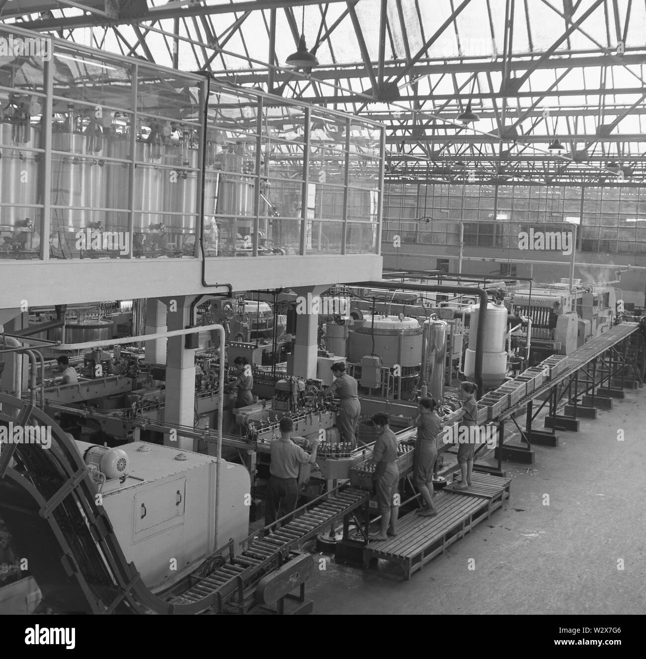 1950er Jahre, historische Erwachsene Männer und Frauen in Overalls, die in der Produktionshalle einer großen Abfüllanlage des Getränkeherstellers Farsons in Malta arbeiten. Stockfoto