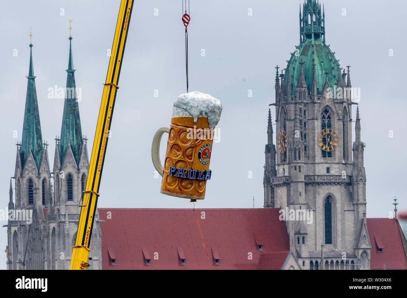 11 Juli 2019, Bayern, München: Ein überdimensionales Bierkrug mit einem Gewicht von rund 1,5 Tonnen ist der Platz von einem Kran auf der Theresienwiese aufgehoben. Im Hintergrund sehen Sie die Sankt Pauls Kirche. Das Oktoberfest beginnt in diesem Jahr am 21. September und endet am 06. Oktober. Foto: Peter Kneffel/dpa Stockfoto