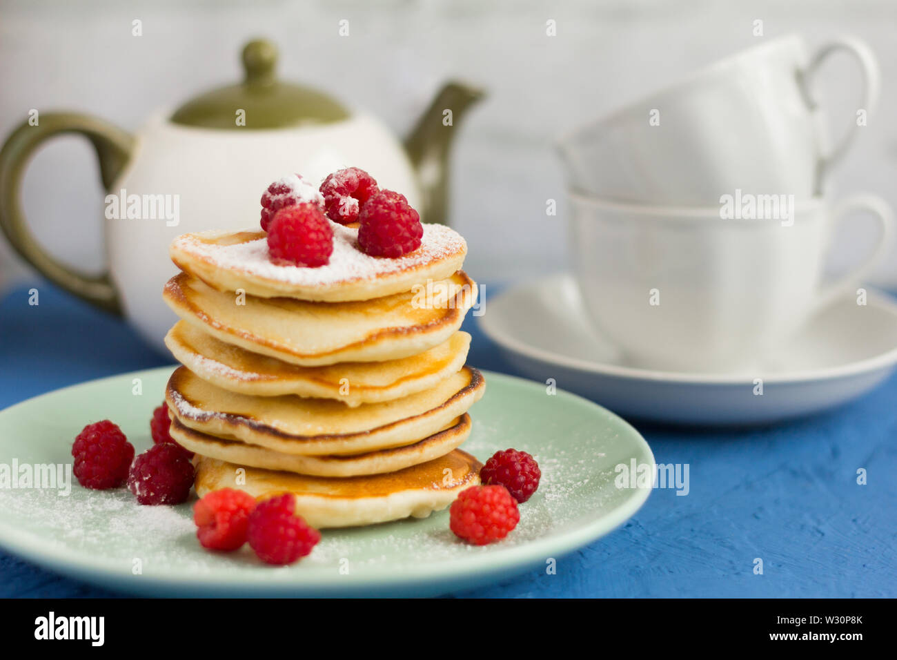 Stapel von gebackenen hausgemachte Pfannkuchen mit Himbeeren, mit Puderzucker und Tee Utensilien im Hintergrund sprengte, selektiver Fokus Stockfoto