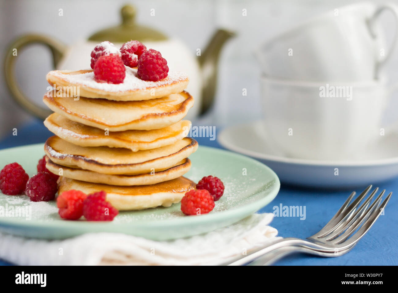 Stapel von gebackenen hausgemachte Pfannkuchen mit Himbeeren, mit Puderzucker und Tee Utensilien im Hintergrund sprengte, selektiver Fokus Stockfoto