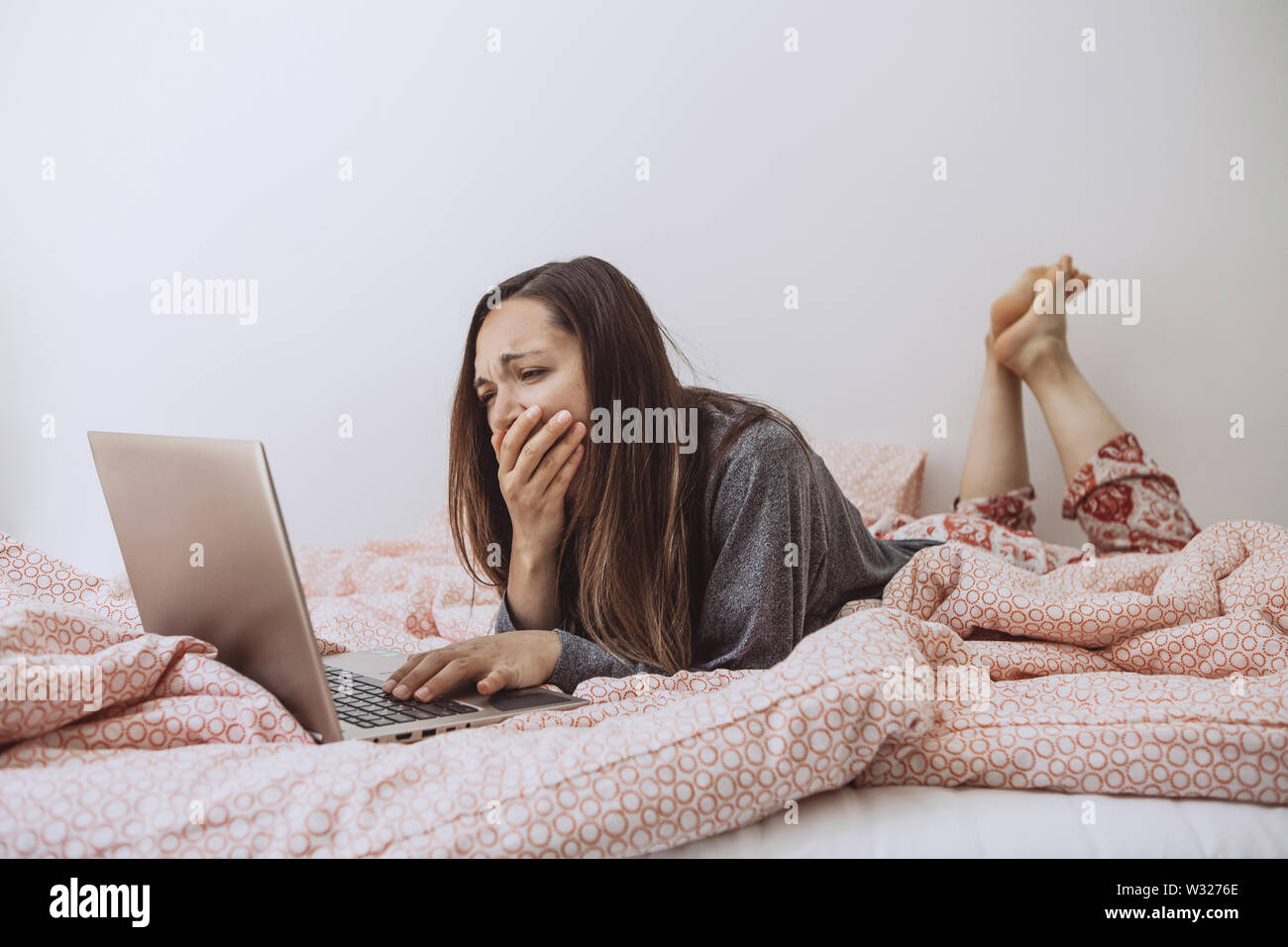 Junge schöne Mädchen genießt einen Laptop im Bett und gähnt. Sie war müde oder wachte früh und schläfrig. Stockfoto