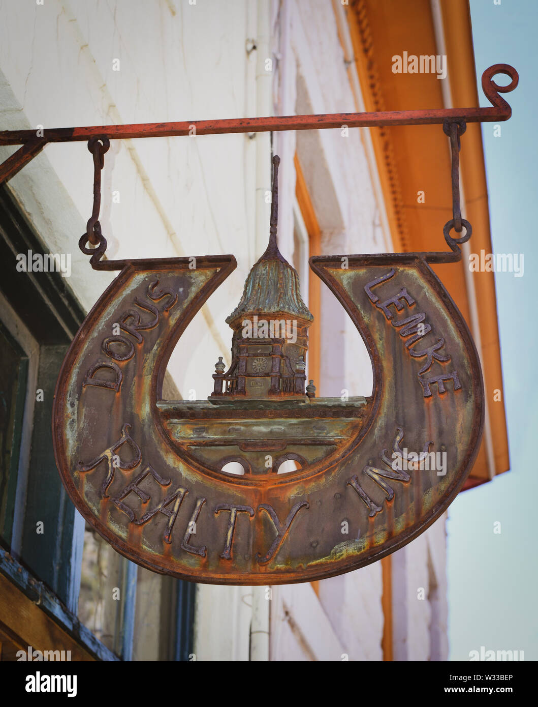 Ein rustikales Kupfer und legierten handgefertigten Geschäft Zeichen für Doris Lemke Realty Inc hängt vom Rahmen Overhead von Office mit lokalen Kirchturm Motiv im Biz Stockfoto