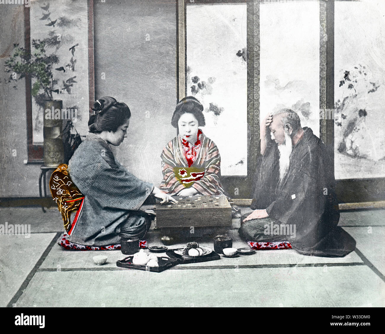 [1890s Japan - Spielen] - Zwei junge Frauen im Kimono und traditionellen Frisuren und ein älterer Mann in einem haori Mantel sind um ein Board für das Go-spiel sitzt. Das Studio ist als Zashiki (Gästezimmer), mit einem Bildschirm, eine ikebana Blumen und blättern Sie in der Rückseite angeordnet. Ein kleines Fach mit einer Teekanne und zwei Tassen kann an der Vorderseite gesehen werden. 19 Vintage Glas schieben. Stockfoto