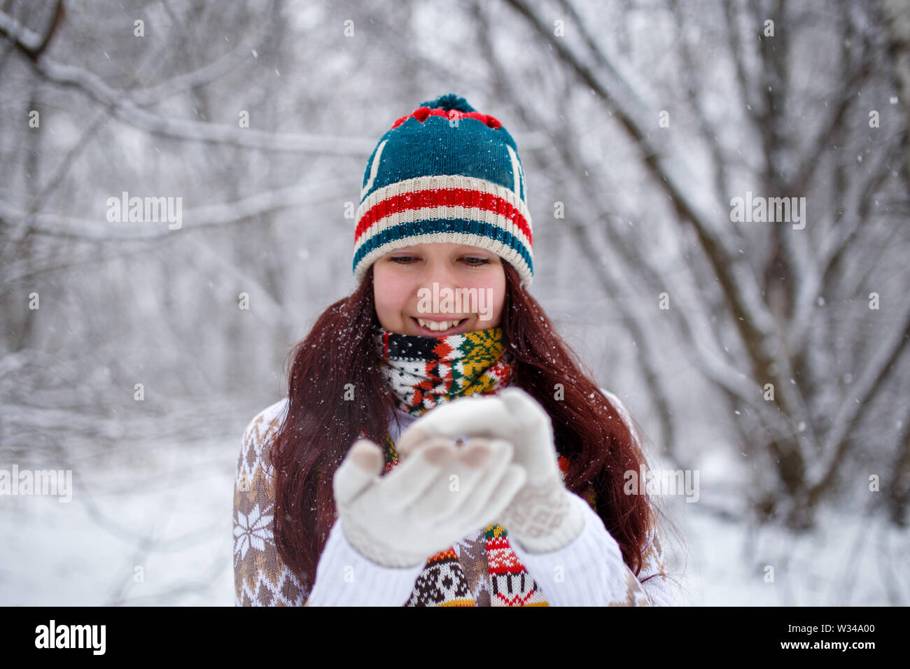 Foto der lächelnde Frau auf gestrickte Mütze und Schal fallende Schneeflocken im Winter Wald bei Tag Stockfoto