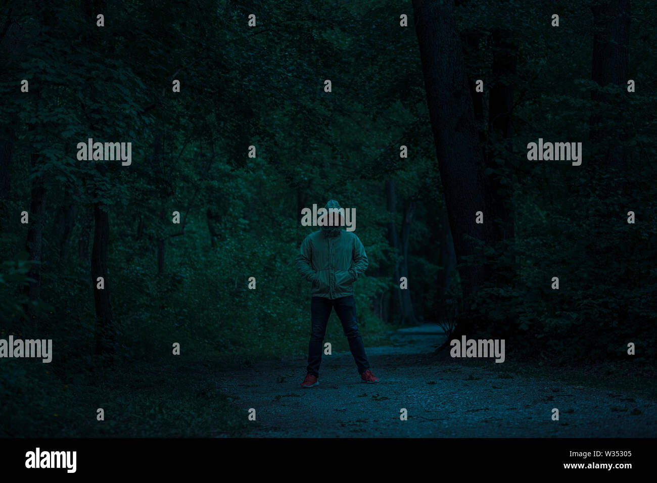 Beängstigend dunkle männliche Figur in einem Hoodie stehend auf dem Pfad in den Wald mit geheimnisvollen Licht hinter ihm kommt, Nacht Horror Szene im Wald Stockfoto