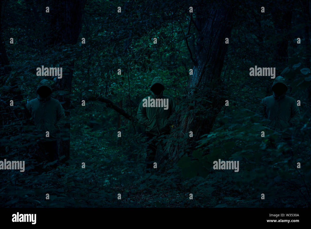 Drei dunkle männliche Figuren in einem Hoodies stehend zwischen Bäumen mit geheimnisvollen Licht hinter Ihnen Nacht Horror Szene im Wald Stockfoto