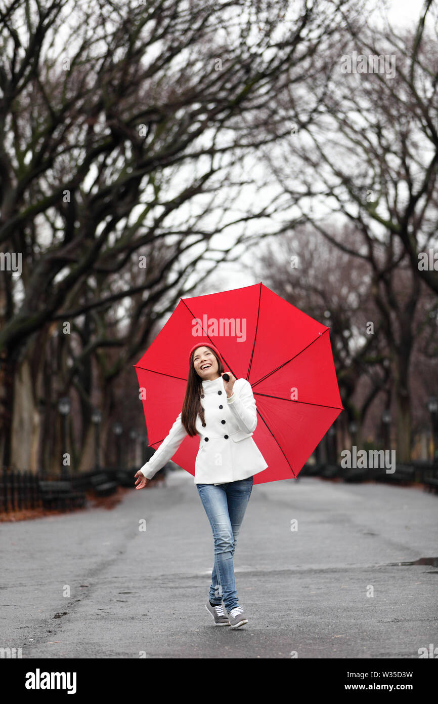 Frau mit Regenschirm im Herbst im Regen oder im Winter. Glücklich lächelnde Mädchen gehen fröhlich mit roten Regenschirm im Central Park, Manhattan, New York City, USA. Stockfoto