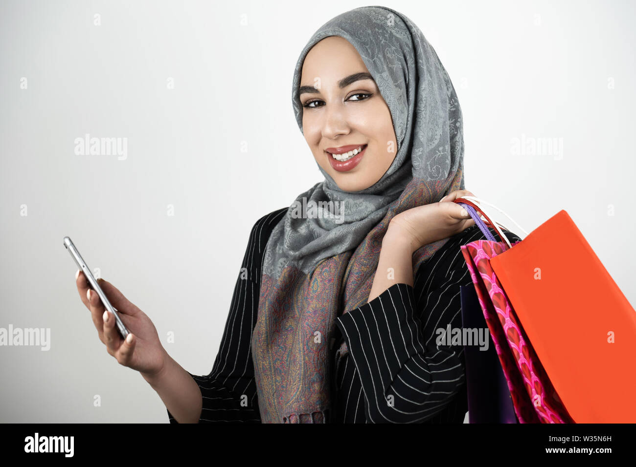 Junge schöne Lächeln Muslimischen business Frau mit Turban hijab Kopftuch holding Smartphone in der einen Hand und Einkaufstaschen in einem anderen isoliert Stockfoto