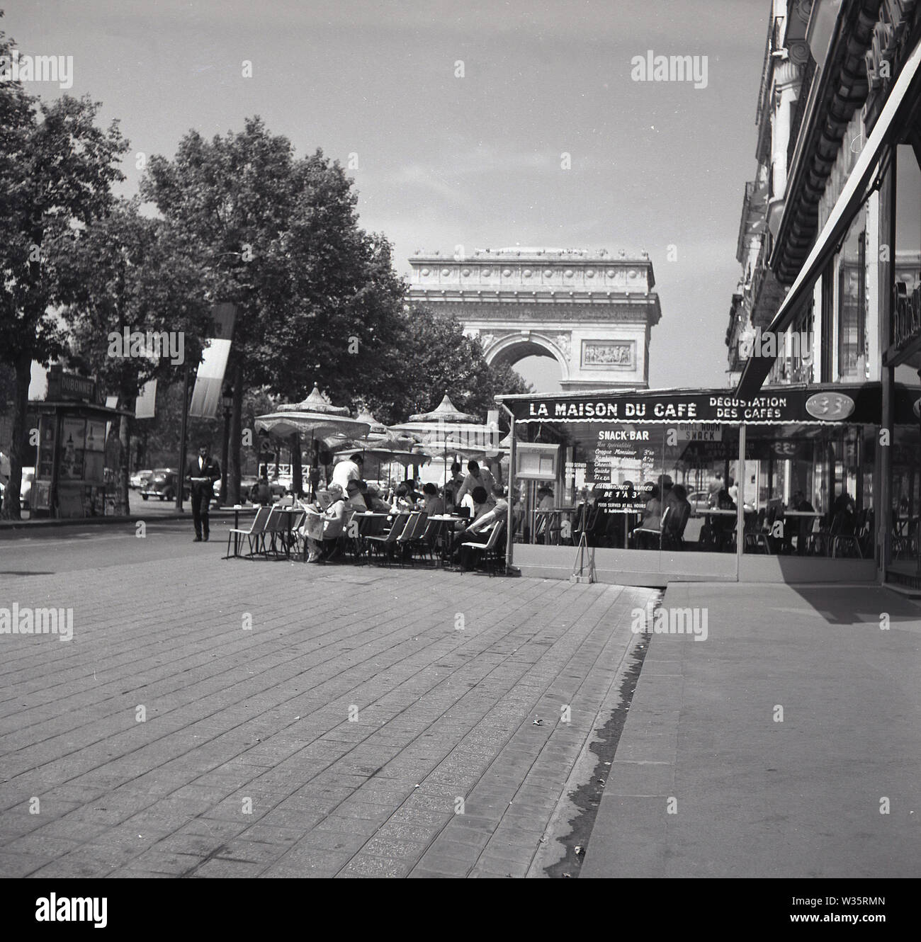 1960er Jahre, historisch, tagsüber und Menschen, die vor dem La Maison du Cafe sitzen, einem Café-Restaurant auf einem Bürgersteig am oberen Ende der Champs-Elysées, mit Blick auf das berühmte parisan-Denkmal, den Arc de Triomphe. Stockfoto