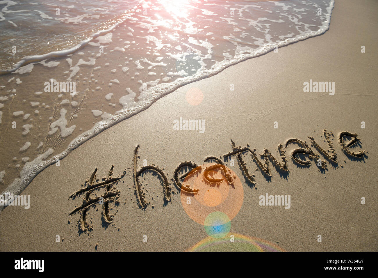 Moderne Nachricht für den Strand Wetter, Hitzewelle, mit einem Social Media - freundliche hashtag in glatten Sand mit Lens Flare geschrieben über einen ankommenden Welle Stockfoto
