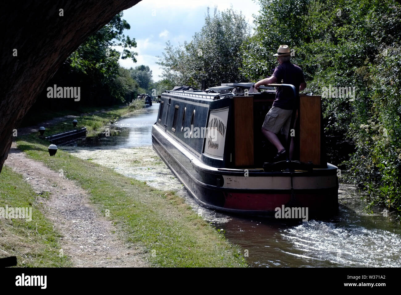 Ein schmales Boot, das von einem Mann mit Sonnenhut auf dem Grand Union Canal direkt nach dem Verlassen einer abgesenkten Schleuse in der Nähe von Aylesbury, Buckinghamshire, gelenkt wird. Stockfoto