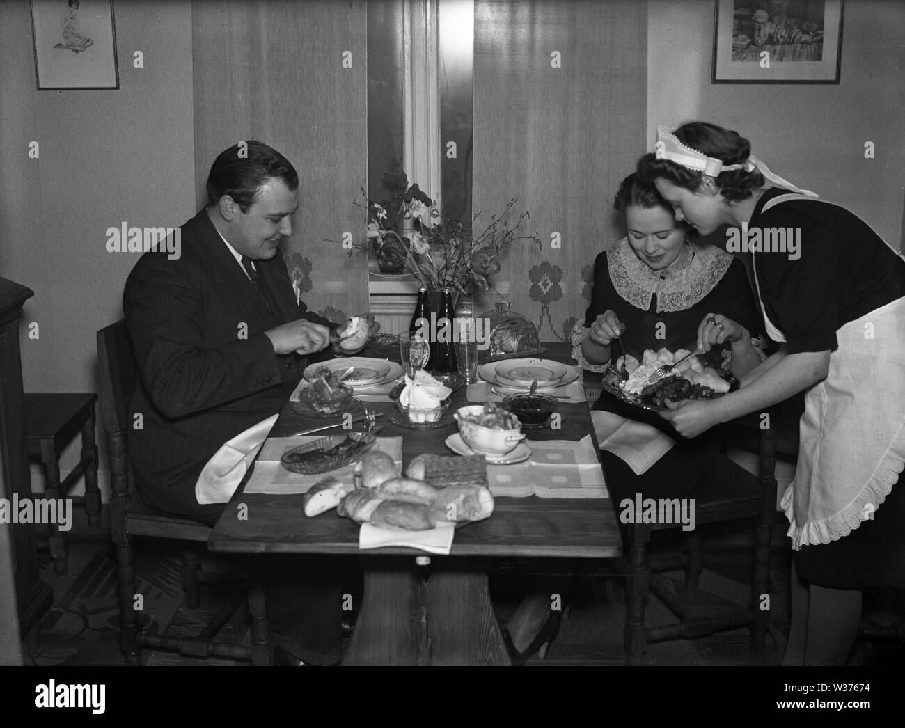Leben in den 1940er Jahren. Eine junge Frau, die als Dienstmädchen ist, das Abendessen serviert, ein Mann und eine Frau am Tisch sitzen. Sie ist ordentlich in einem weißen Schürze gekleidet. Schweden 1940. Kristoffersson ref 55-7 Stockfoto